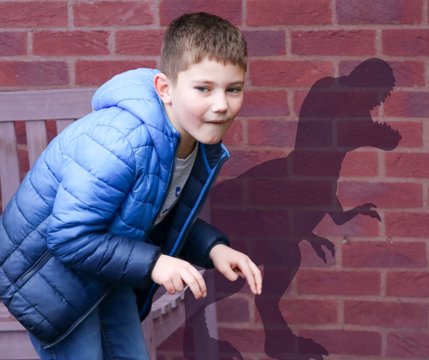 A young boy aged 8 pretending to be a dinosaur. He is hunched over with t-rex hands, and his shadow is cast on a wall behind him depicting a roaring t-rex