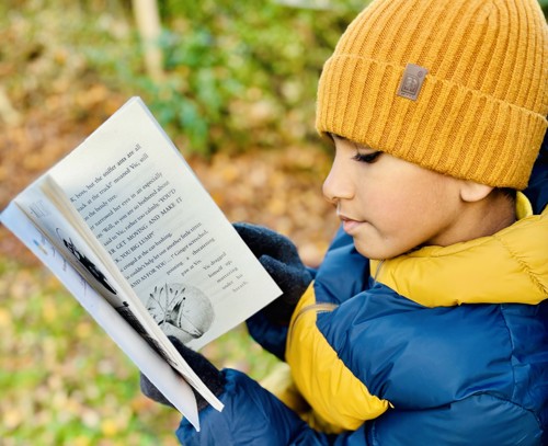 a young boy reading outdoors wearing a woolly hat and gloves and a puffer coat